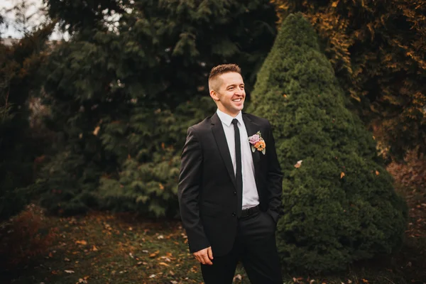 Beautiful stylish young groom in black wedding suit white jacket shirt tie, standing outdoors in the woods, on a natural background