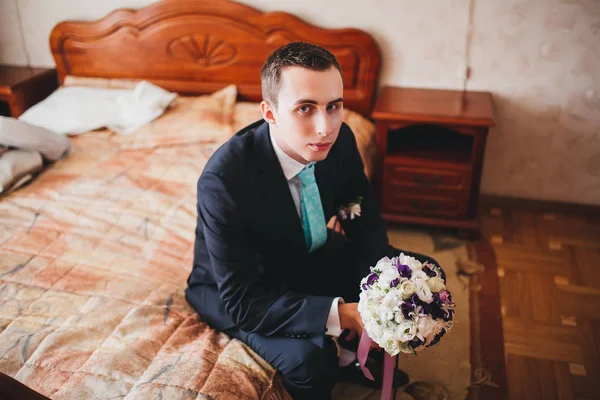 Gorgeous smiling groom. Handsome man in a suite with a buttonhole sitting on window sill indoors