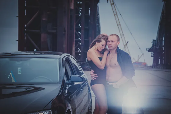 Beautiful blonde girl standing sexy on the unfinished bridge, with the light hair men and car, in black corset on a windy day, grey clouds, holding each other