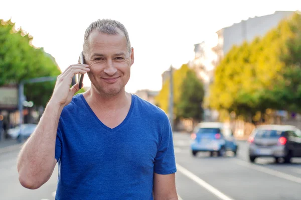 Handsome man holding mobile phone, using smartphone, making a call, talking on the phone, standing on sunny street with transport traffic on the background
