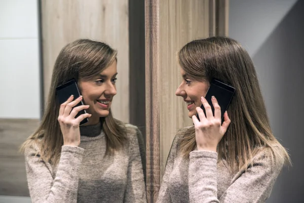 Portrait of a happy woman looking at her reflection in the mirror and talking on the phone. Beautiful smiling young woman talking on phone