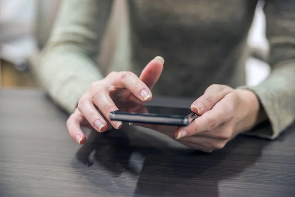 Woman holding a smartphone on the wooden table. Close-up hands of the girl, sitting at the wooden table, in one hand is smartphone. Businesswoman surfing the Internet on smartphone.