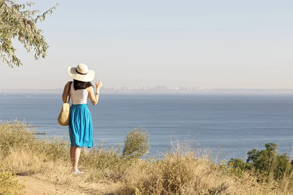 Young woman (brunette) in a blue skirt and hat looks at sea.