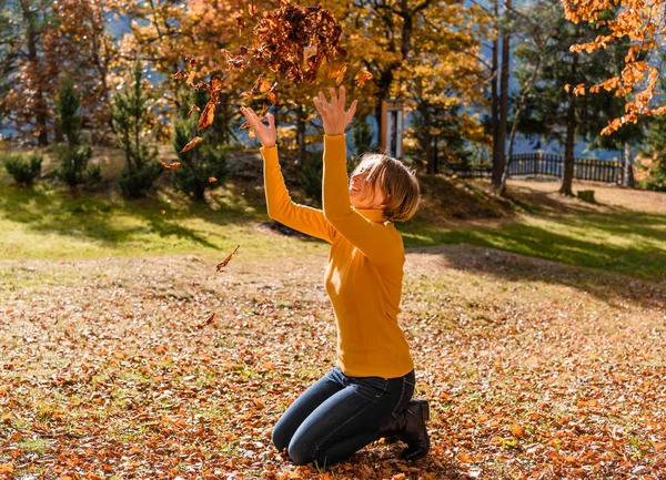 Happiness carefree. Woman has a fun in autumn park throwing leaves up in the air with arms raised up. Beautiful girl smiling and playing in colorful forest foliage outdoor.