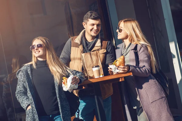 Group of cheerful young people talking, drinking coffee and eating croissant in cafe outdoor on sunny day. Concept togetherness, friendship, communication.