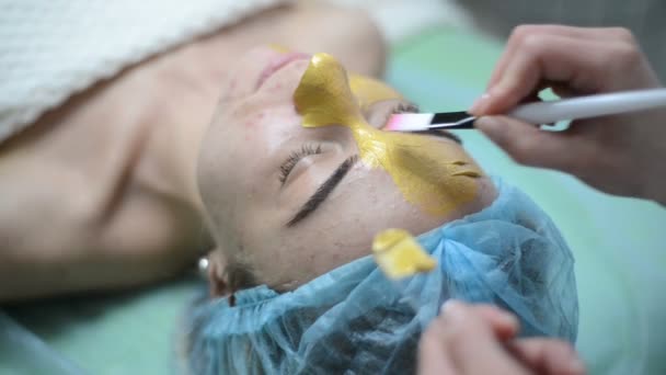 Cosmetologist εφαρμόζοντας χρυσό του προσώπου μάσκα στο πρόσωπο, γυναίκα που έχει αισθητική θεραπεία στο spa. Επαγγελματίας αισθητικός με γυναίκα στο σαλόνι ομορφιάς. Επεμβάσεις στο δέρμα καθαρισμού κοσμετολογία. — Αρχείο Βίντεο