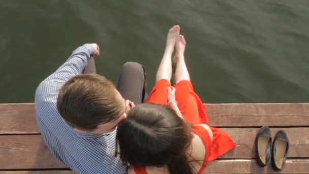 Pareja feliz sentada en el muelle en la orilla del lago sobre el fondo de la puesta del sol — Vídeos de Stock