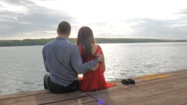 Casal feliz sentado no cais na margem do lago contra o pano de fundo do pôr do sol — Vídeo de Stock