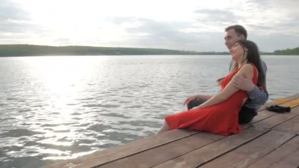 Pareja feliz sentada en el muelle en la orilla del lago sobre el fondo de la puesta del sol — Vídeo de stock