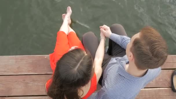 Pareja feliz sentada en el muelle en la orilla del lago sobre el fondo de la puesta del sol — Vídeo de stock