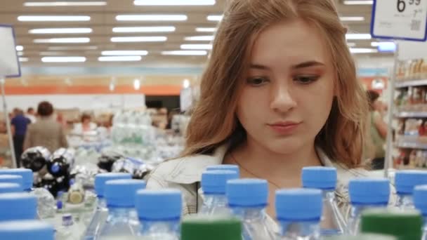 Jovem mulher fazendo compras no supermercado. Ela compra escolher uma garrafa de água mineral e colocá-lo em seu cesto de compras. Compras na mercearia — Vídeo de Stock