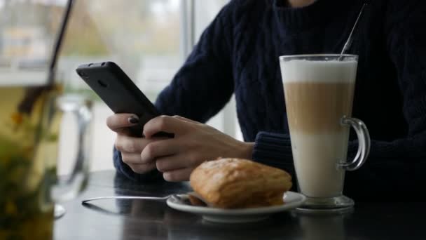 Młoda ładna kobieta przy użyciu smartfona w kawiarni, wypić kawę w Puchar, śniadanie na słodko, szczęśliwy twarz, odkryty hipster portret, Moda dziewczyna, stół, Kawa latte smaczny słodki, telefon — Wideo stockowe