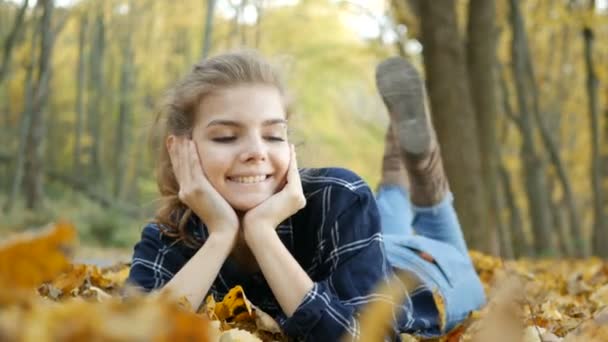 Молодая красивая девушка в синих джинсах лежит на желтых листьях, в осеннем парке — стоковое видео