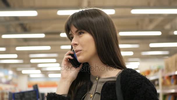 Mujer sonriente haciendo compras en el supermercado y teniendo una llamada telefónica con su teléfono inteligente — Vídeo de stock