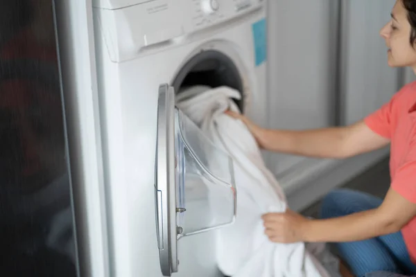 A girl loads dirty laundry into a washing machine while sitting on the floor in an apartment. Laundry day, housework.