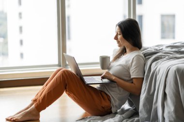 Güzel bir genç esmer kız dizüstü bilgisayarda çalışıyor ve kahve içiyor, panoramik pencerenin yanındaki yatağın yanında yüksek kattan manzaralı yerde oturuyor. Modern iç mimari..