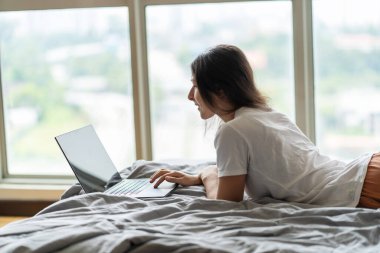 Güzel bir genç esmer kız, panoramik bir pencerenin yanındaki yatağın üstünde üst kattan güzel bir manzarayla çalışırken dizüstü bilgisayarda çalışıyor. Modern iç tasarımı çok şık. Rahat bir iş yeri. İnternet Alışverişi