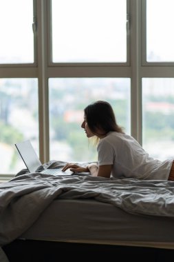 Güzel bir genç esmer kız, panoramik bir pencerenin yanındaki yatağın üstünde üst kattan güzel bir manzarayla çalışırken dizüstü bilgisayarda çalışıyor. Rahat bir iş yeri. İnternetten alışveriş.