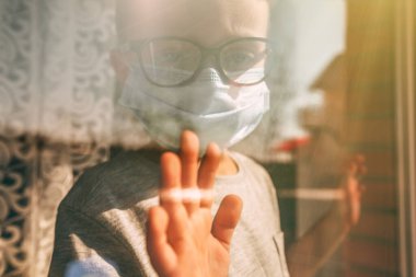 Virüs yüzünden karantinada olan bir çocuk maske takıp güneşli bir günde pencereden dışarı bakar.