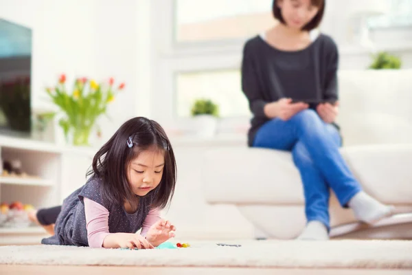 Roztomilá holčička hraje na koberec a matka sedí s tabulkou — Stock fotografie