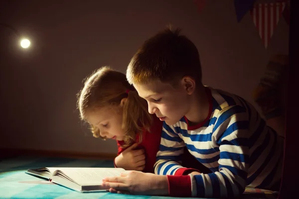 Twee gelukkige broers en zussen lezen boek bij duisternis met zaklamp bij h — Stockfoto