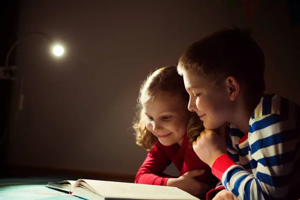 Twee gelukkige broers en zussen lezen boek bij duisternis met zaklamp bij h — Stockfoto