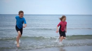 Neoprene mayo giymiş, kumsalda koşan, dalgalarla ve kumla oynayan oldukça mutlu bir kızın yavaş çekim videosu.