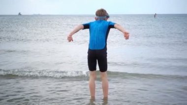 Neoprene mayo giymiş, kumsalda koşan, dalgalarla ve kumla oynayan mutlu bir çocuğun yavaş çekim videosu.