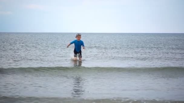 ビーチで実行されているネオプレン水着で幸せな男の子のスローモーションビデオと波と砂で遊ぶ — ストック動画
