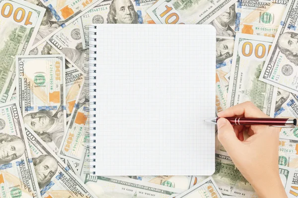 Hand with pen over blank notebook on background of dollar bills and coins — Stock Photo, Image