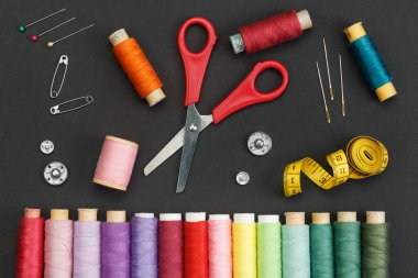 Siyah zemin üzerinde renkli iplik bobinleri, makas, düğmeler, iğneler, iğne ve bir terzi sayacı.