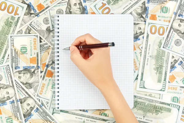 Hand with pen over blank note book on background of dollar bills and coins — Stock Photo, Image