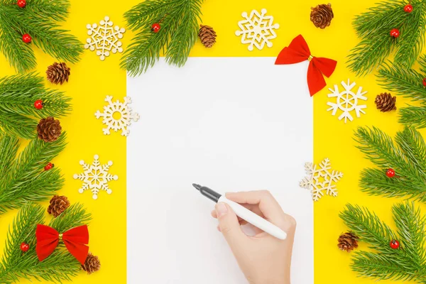 Mano con marcador sobre un papel blanco en blanco, borde de Navidad con ramas de abeto, ceniza de montaña, conos en un amarillo — Foto de Stock
