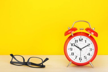Red alarm clock and glasses on a wooden table against a yellow background. clipart