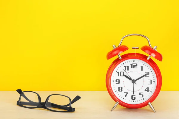 Red alarm clock and glasses on a wooden table against a yellow background. — Stock Photo, Image