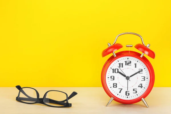 Reloj despertador rojo y vasos sobre una mesa de madera sobre un fondo amarillo . Fotos de stock libres de derechos