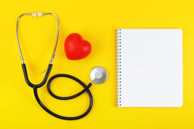 Kırmızı kalp ve steteskop. Sarı arka planda boş not defteri var..