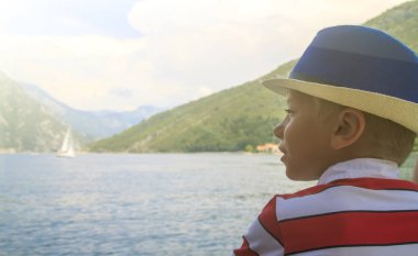 kotor Körfezi boyunca bir feribot üzerinde mavi şapkalı çocuk yelken ve coşkuyla dağlara ve bir yelkenli tekne ye uzaklık içine bakar
