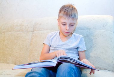 Küçük çocuk evde bej koltukta oturmuş kitap okuyor ve içindekilerle çok ilgileniyor.