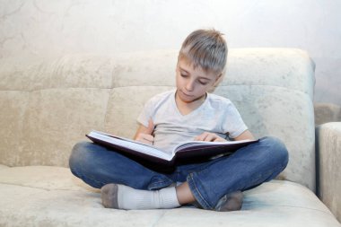 Küçük çocuk evde bej koltukta oturmuş kitap okuyor ve içindekilerle çok ilgileniyor.