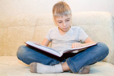 Küçük çocuk evde bej koltukta oturmuş kitap okuyor ve içindekilerle çok ilgileniyor.