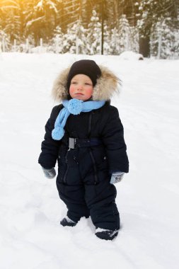 Sakin, düşünceli küçük çocuk, arkasında sessiz bir kış ormanı olan karla kaplı yerde duruyor.