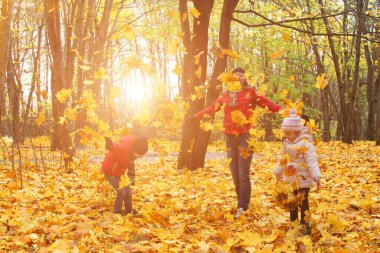 Mutlu bir aile güneşli parkta yürüyor ve turuncu akçaağaç yaprakları fırlatıyor. Anne ve çocuklar, sonbahar havasının tadını çıkarıyorlar.