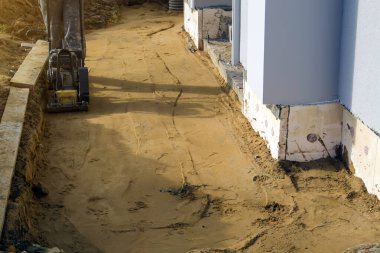 İşçi, binanın etrafındaki kumu özel çalışma aracı (tamping makinesi veya titreşim plakası) ile sıkıştırır)