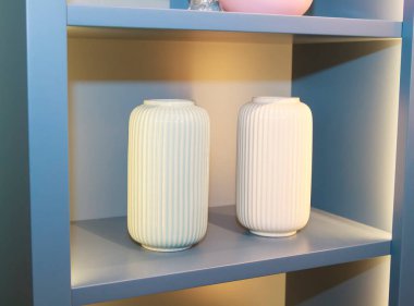 Mavi bir rafta iki beyaz seramik vazo.