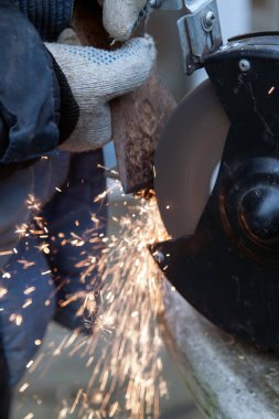 Eldiven ve mavi ceketin ustası dışarıda elektrikli bileyicinin baltasını keskinleştiriyor. Farklı yönlerde uçuşan kıvılcımlarla keskinleştirme işlemi