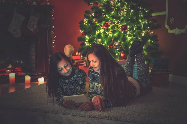 Jóvenes amigas tumbadas en la alfombra junto al árbol de Navidad y mirando la tableta. Hermosa habitación de año nuevo interior —  Fotos de Stock