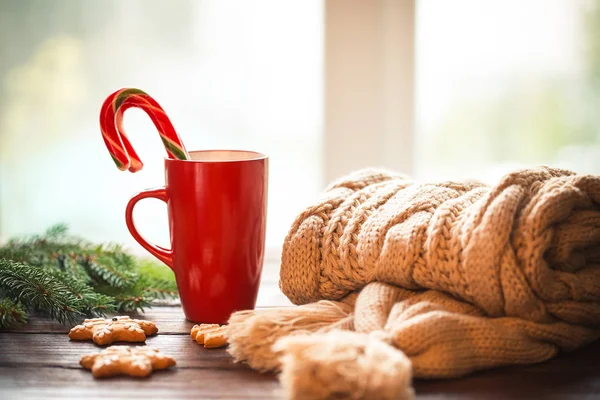 Christmas gingerbread, candy, coffee in red cup, knitted scarf, cones  and christmas tree branches on wooden table on a frosty winter day window background. Home cozy holidays.