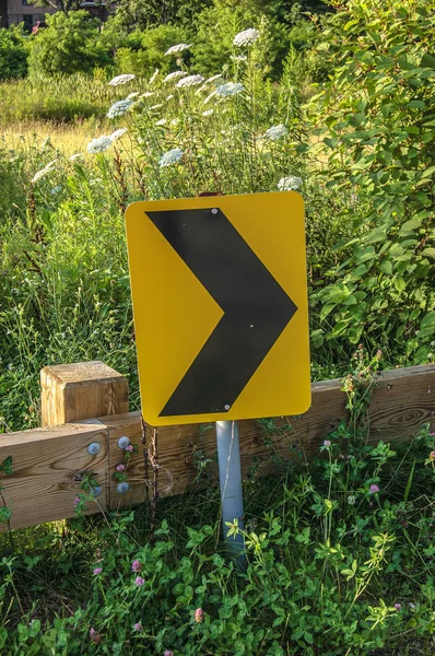 Дорожный знак поворота на обочине перед зелеными растениями и деревянным забором — стоковое фото