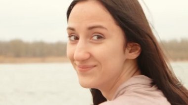Nehri yakınında kahverengi saçlı kız closeup çekim geri bak. Uzun saçlı esmer kız açık havada riverside su yüzeyi ve uzak riverbank ağaçta önünde poz. Vintage Renk tonlu